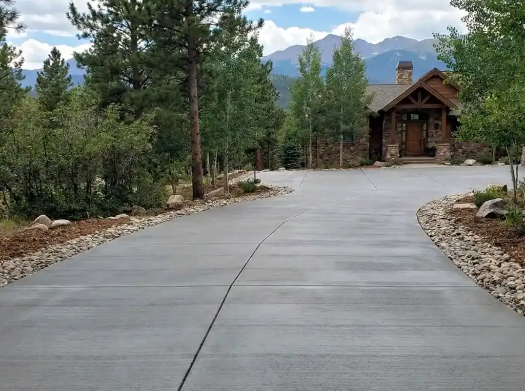 Gray broom-finished concrete driveway with smooth control joints leading to a mountain home surrounded by pine trees