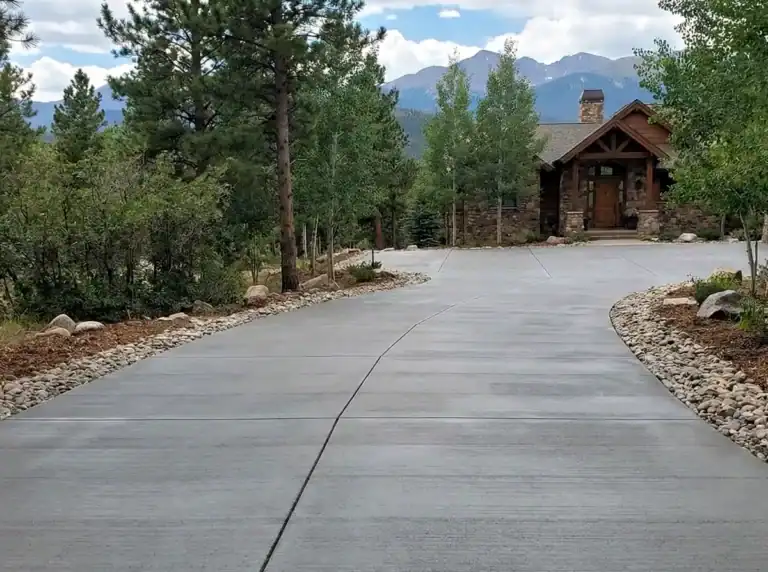 Gray broom-finished concrete driveway with smooth control joints leading to a mountain home surrounded by pine trees